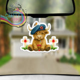 A brown highland cow car air freshener wearing a blue beret with a red poppy between two white crosses with poppies