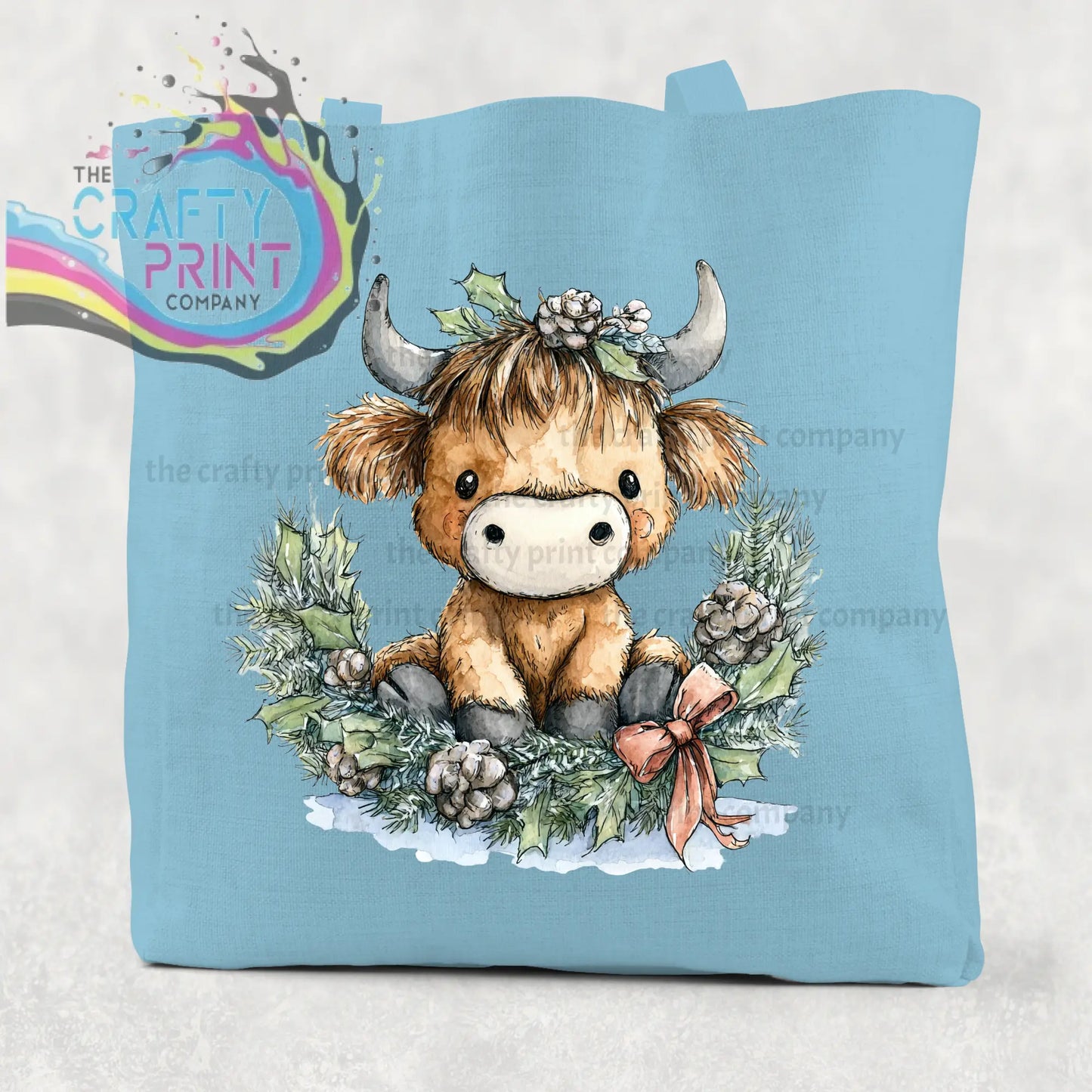 Light blue cotton tote bag featuring a brown highland cow wreath with holly and pinecones in a festive design