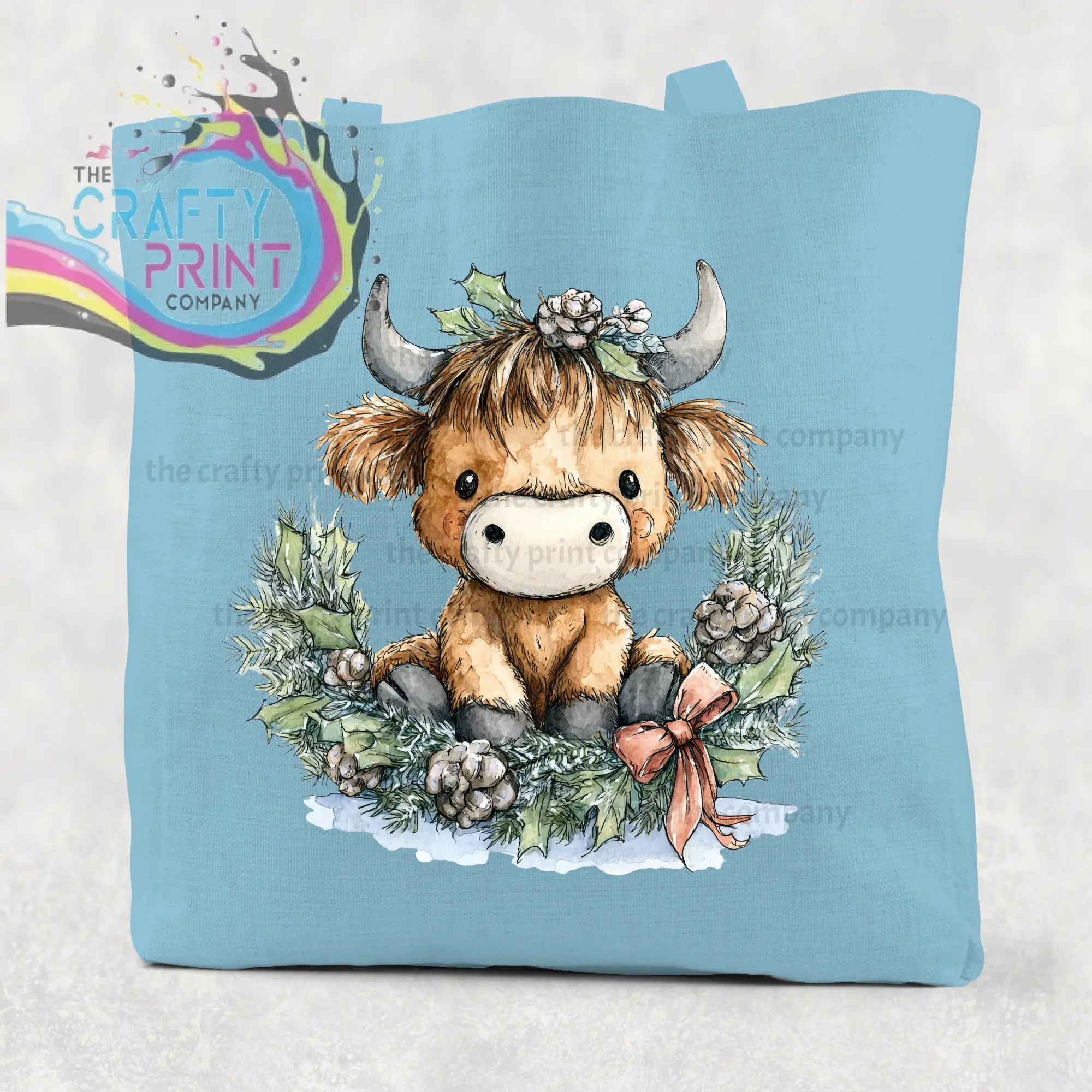 Light blue cotton tote bag featuring a brown highland cow wreath with holly and pinecones in a festive design
